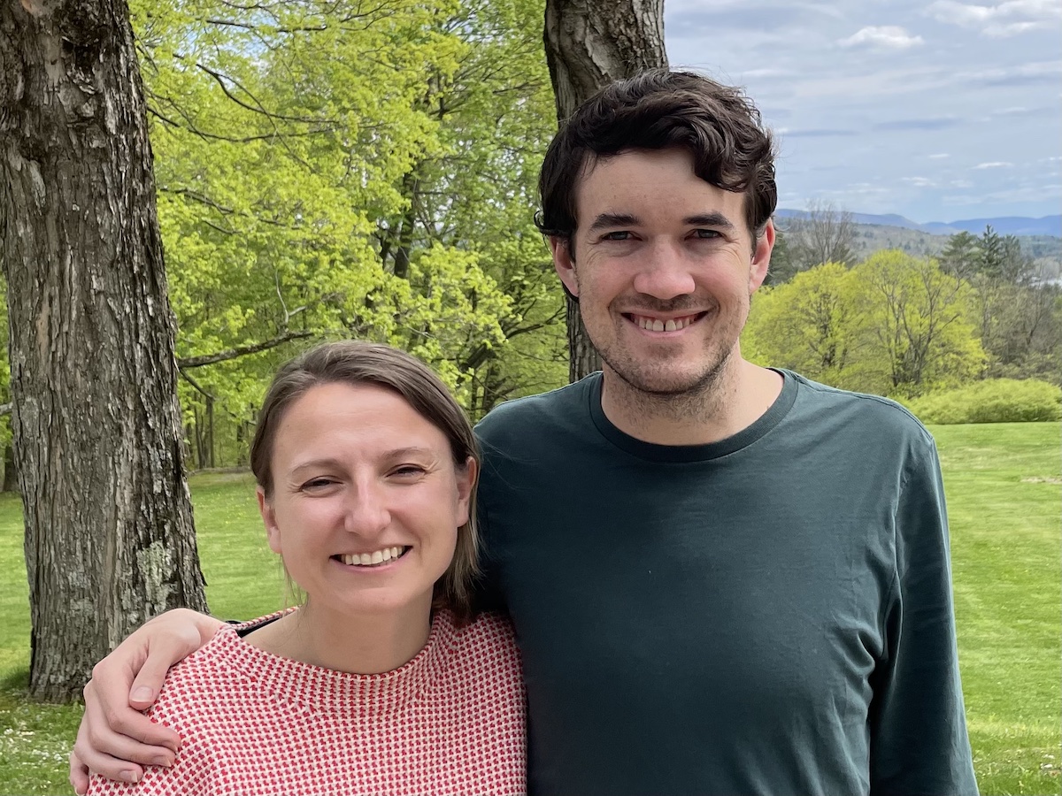 Claire and Toph, smiling at the camera, Toph’s arm lovingly around Claire’s shoulder, standing at the top of the Apple Tree hill in front of the unseen old carriage house, a beautiful verdant backdrop with layered mountains and just a hint of the Stockbridge Bowl visible at the far right